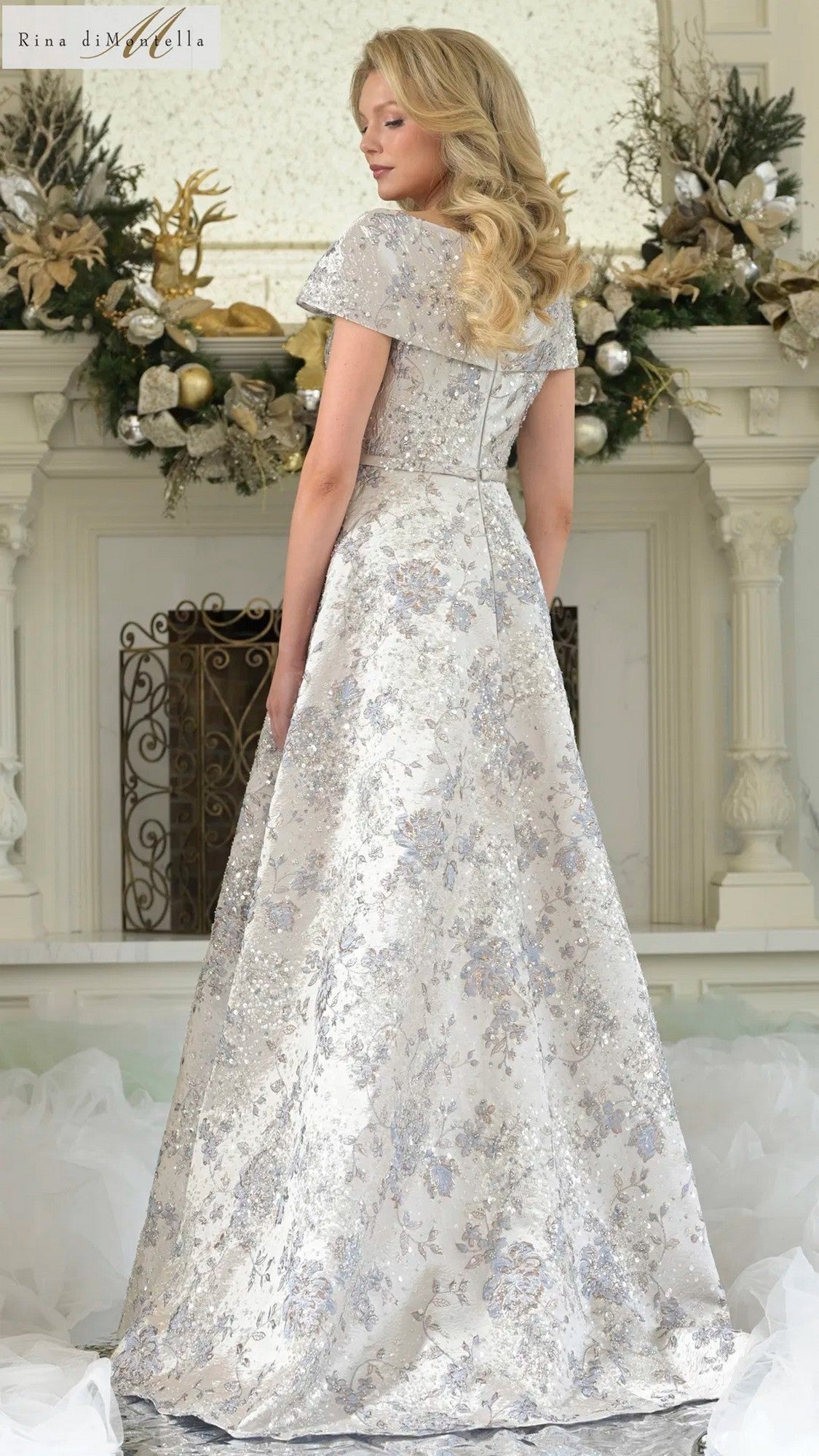 A woman with long blonde hair models the Rina Di Montella RD3312 silver floral jacquard long formal dress, standing with her back to the camera before an ornate fireplace with gold accents.